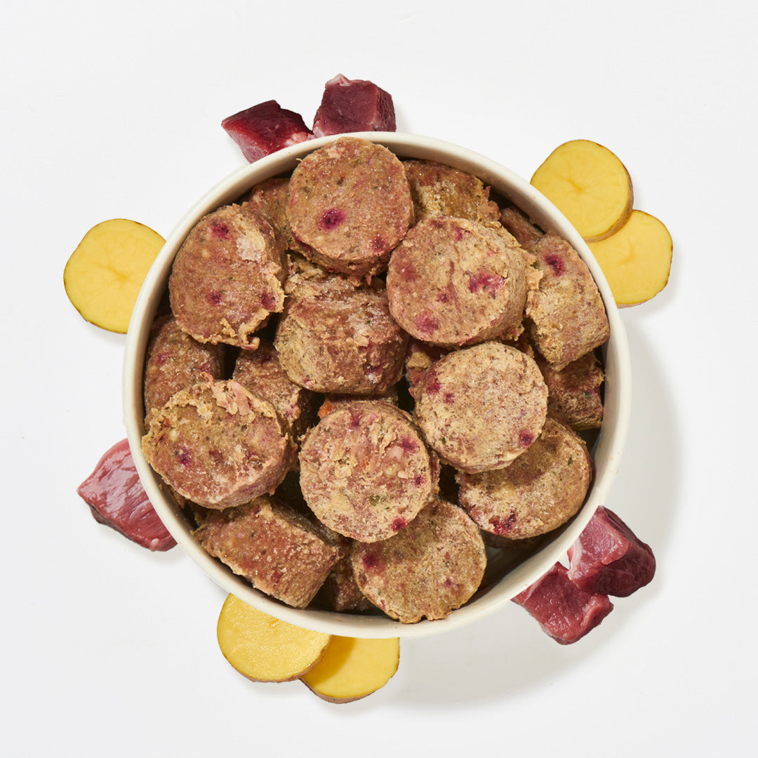 Schüssel mit All-in-One Nuggets Rind & Lamm Katzenfutter umgeben von rohem Fleisch und Gemüse, hochwertige, getreidefreie Komplettmahlzeit.