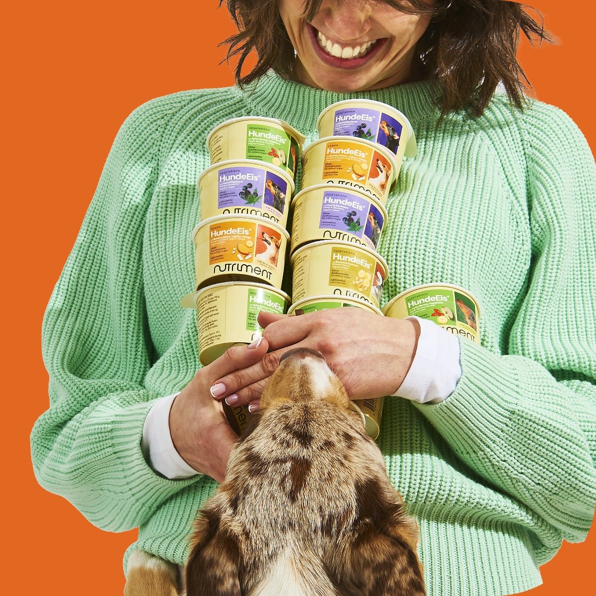 Frau hält Nutriment HundeEis Becher, Hund schnuppert veganem Hundeeis Banane Kokos-Joghurt Mango